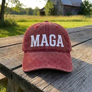 May include: A red baseball cap with a distressed, washed-out finish. The front of the cap features the word MAGA embroidered in bold, white, capital letters. The cap has a structured crown and a curved brim, showcasing a classic six-panel design.