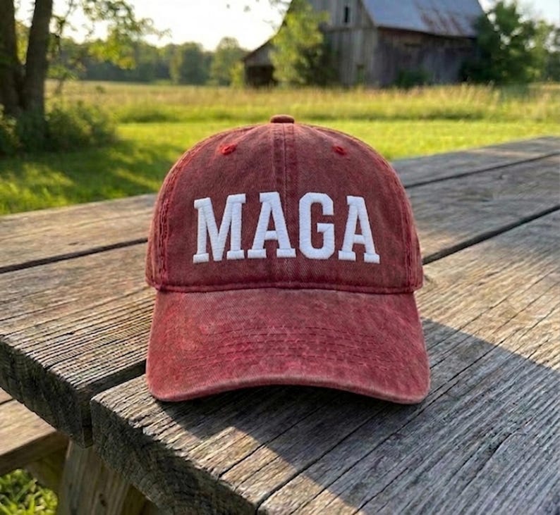 May include: A red baseball cap with a distressed, washed-out finish. The front of the cap features the word MAGA embroidered in bold, white, capital letters. The cap has a structured crown and a curved brim, showcasing a classic six-panel design.