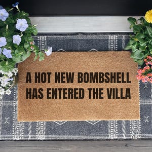 May include: A rectangular tan coir doormat featuring the black text A HOT NEW BOMBSHELL HAS ENTERED THE VILLA. The mat is layered over a dark gray and white plaid rug, positioned between two potted plants with blue, white, yellow, and pink flowers.