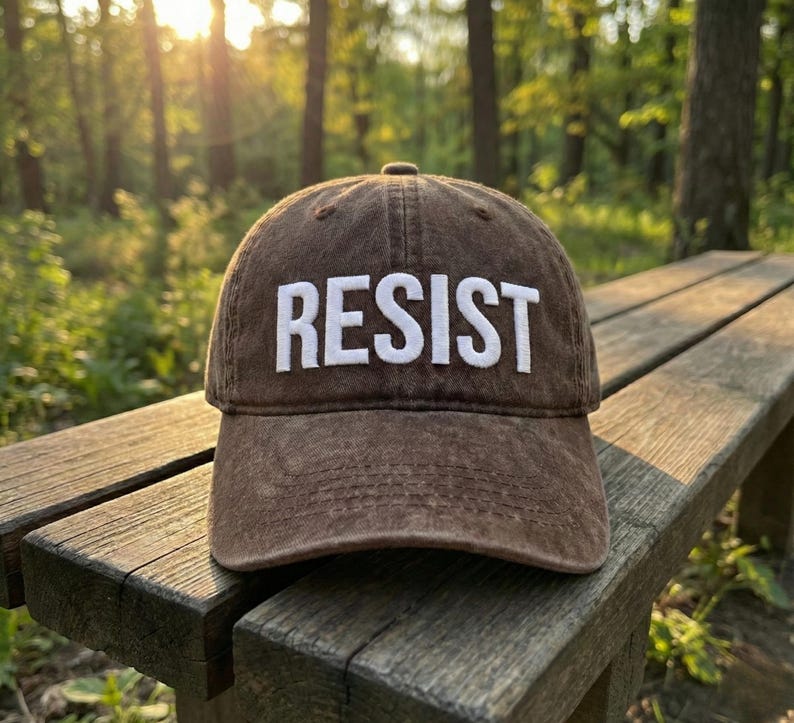 May include: A brown baseball cap with the word "RESIST" embroidered in white block letters. The cap is sitting on a wooden bench in a park setting, with trees and sunlight in the background.