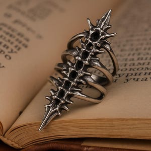 May include: A silver-toned ring with a unique, spiky design. The ring features a series of pointed projections and embedded black stones, creating a bold and edgy aesthetic. The ring's design wraps around the finger, offering a distinctive look. The ring is set against a backdrop of an open book.