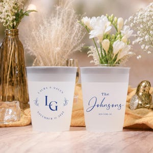 May include: Two frosted plastic cups with dark blue text and floral accents. One cup reads "Laura & Galen" with initials and a date. The other cup says "The Johnsons" and a location. Wedding or party theme.