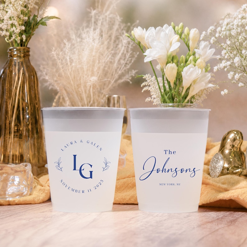 May include: Two frosted plastic cups with dark blue text and floral accents. One cup reads "Laura & Galen" with initials and a date. The other cup says "The Johnsons" and a location. Wedding or party theme.