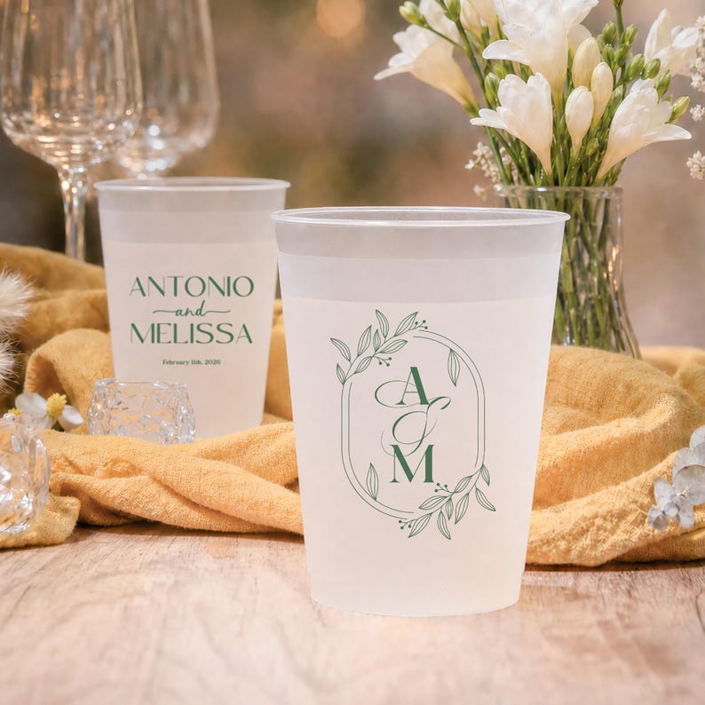 May include: Frosted plastic cups with custom green text and floral designs. One cup reads "Antonio and Melissa" with a date, while another features the initials "A M" inside a decorative frame. Wedding or event theme.