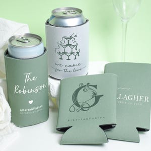 May include: Sage green can coolers with personalized text and designs. One cooler says "The Robinson" with a heart and date. Another says "we came for the love" with a martini glass design. Other coolers have initials and names.