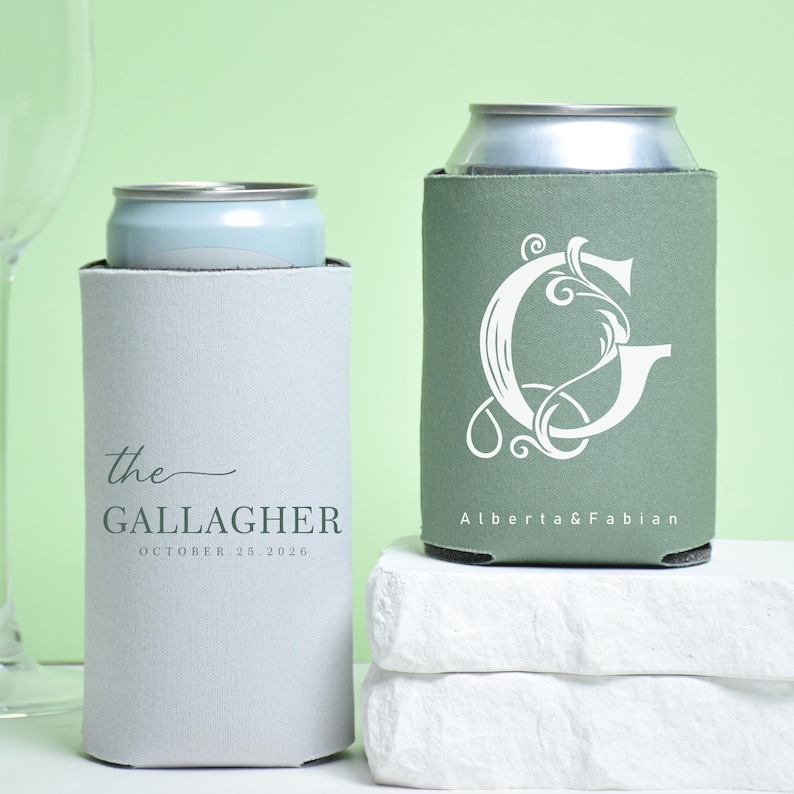 May include: Two can coolers, one light gray and one sage green, each holding a silver can. The gray cooler has the text "the GALLAGHER OCTOBER 25 2026". The green cooler features a white ornate letter "G" and the names "Alberta & Fabian".