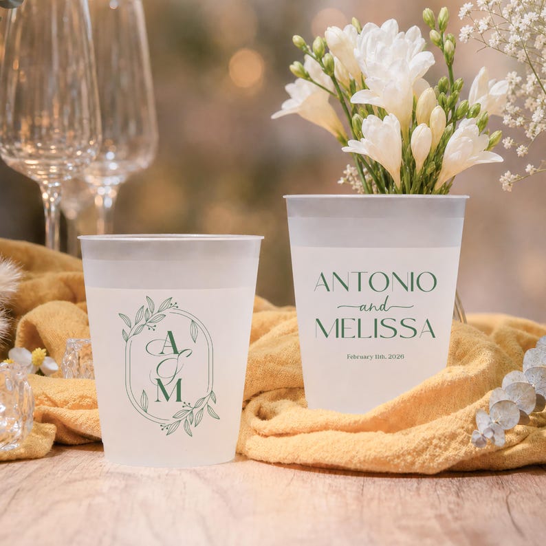 May include: Two frosted plastic cups with white rims. One cup features the initials "A" and "M" inside a green floral design. The other cup has the names "ANTONIO and MELISSA" and the date "February 8th, 2026" in green.