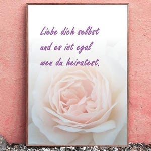 Könnte beinhalten: Ein gerahmter Druck mit einem Bild einer hellrosa Rose und dem deutschen Text "Liebe dich selbst und es ist egal wen du heiratest" in Lila. Der Rahmen ist braun, vor einer rosa Wand. Der Druck liegt auf kleinen Steinen.