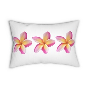 Puede incluir: Cojín rectangular blanco con tres flores de plumeria estilo acuarela. Las flores tienen pétalos rosas con centros amarillos. La almohada es un artículo decorativo, probablemente para una cama o un sofá.