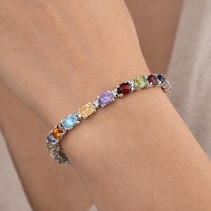May include: A silver bracelet featuring a series of oval-cut gemstones in a rainbow of colors. The gemstones are set in a row, creating a vibrant and eye-catching design. The bracelet is worn on a wrist.