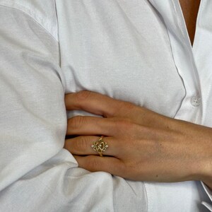 May include: A gold-colored ring with an intricate floral design, featuring small, clear gemstones. The ring is worn on a hand, with a white button-down shirt in the background. The ring's design includes multiple loops and small, sparkling accents.