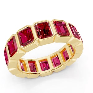 Op de afbeelding: Een gouden ring met een reeks rechthoekige, levendige rode edelstenen. De edelstenen zijn in een herhalend patroon rond de band gezet. De ring heeft een gepolijst, reflecterend oppervlak en de edelstenen zijn dieprood.
