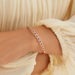 Pode incluir: Uma pulseira dourada com uma fileira de pedras preciosas vermelhas redondas. A pulseira é usada em um pulso, contra um fundo de tecido creme. As pedras preciosas são dispostas em um padrão repetitivo.