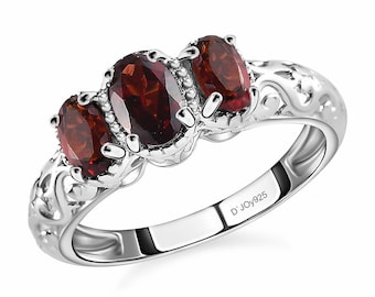 May include: A silver ring featuring three oval-cut, deep red gemstones. The ring has an ornate, swirling design on the band and is marked with "D'JOY925". The gemstones are set in a prong setting.
