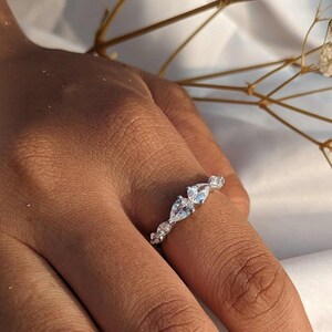 May include: A silver ring with a series of clear, pear-shaped and round gemstones. The ring is on a person's finger, with a white background and dried flowers in the background.