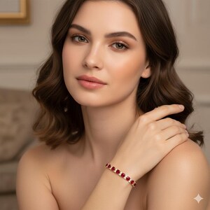 May include: A gold bracelet with alternating red gemstones and clear, smaller stones. The bracelet is worn on a wrist. The gemstones are rectangular and set in a gold band. The background is blurred.