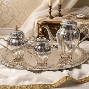 May include: A silver tea service set consisting of a large teapot, a sugar bowl, and a creamer, all featuring vertical fluted panels, ornate floral engravings, and small finials on the lids. The pieces are arranged on a large, oval silver tray.