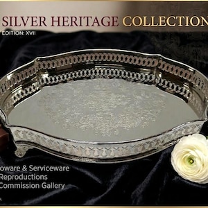 Peut inclure: Un plateau de service argenté avec des détails ornés, de la "Collection Silver Heritage". Le plateau a un bord décoratif et repose sur de petits pieds. Un coffret cadeau avec un ruban doré et une fleur blanche sont également présents.