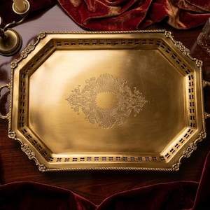 May include: An octagonal brass serving tray featuring a central etched floral pattern, a pierced lattice border, and two ornate handles. The tray has a polished metallic finish with decorative embossed details along the outer rim and edges.