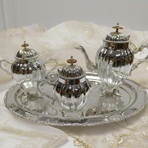 May include: A silver tea set, including a teapot, sugar bowl, and creamer, sits on a matching tray. The set has ornate detailing and is displayed on a white and gold patterned surface. The teapot has a height of approximately 7 inches.