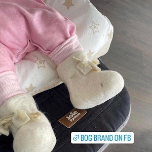 May include: A baby wearing light pink clothing and off-white felt booties with bow accents. The booties are on a black and white patterned surface. The image includes the text "BOG BRAND ON FB".