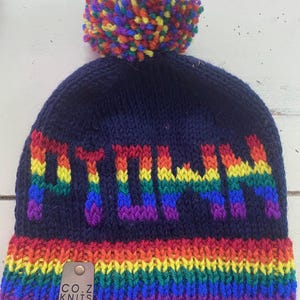 May include: A navy blue knit beanie with a rainbow pom-pom and rainbow striped brim. The word "PTOWN" is knit in rainbow colors across the front. A small leather tag reads "CO.Z KNITS".