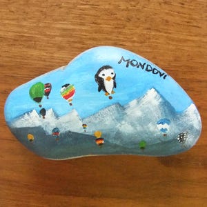 Può includere: Sasso dipinto a mano con sfondo di cielo blu, montagne e mongolfiere colorate. Un pinguino è raffigurato nel cielo, con la scritta "Mondovi" sopra. Il sasso è su una superficie di legno.