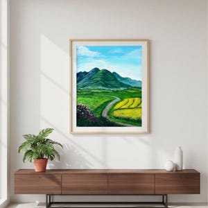 Può includere: Un dipinto di paesaggio incorniciato raffigura una strada tortuosa attraverso campi verdi e gialli, che conduce a una catena montuosa sotto un cielo blu. L'opera d'arte è in una cornice di legno chiaro, aggiungendo un tocco naturale alla scena.
