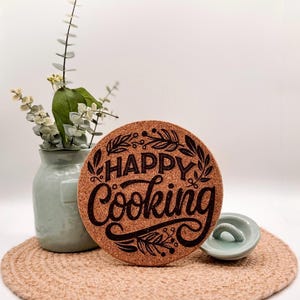 Könnte beinhalten: Ein runder Topfuntersetzer aus Kork mit den Worten "HAPPY Cooking" in schwarzer Schrift und dekorativen Blättern. Der Untersetzer liegt auf einem gewebten Platzset, neben einer kleinen grünen Vase mit Grün und einer kleinen, hellgrünen Schale.