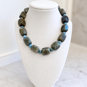 May include: A necklace featuring large, rectangular, polished grey-brown stones with iridescent flecks, alternating with small, round, light blue beads. The stones have a smooth, rounded finish and are arranged in a single strand circular pattern.