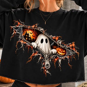 May include: A black long-sleeve sweatshirt featuring a graphic print of a zipper opening to reveal a white ghost, orange pumpkins, and a haunted house scene. The design includes spiderwebs and a zipper pull shaped like a jack-o'-lantern face.