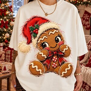 May include: A white short-sleeved t-shirt featuring a large graphic of a gingerbread person wearing a red Santa hat with a white pom-pom, a red and green plaid bow tie, and two peppermint candy buttons on the chest.