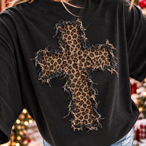 May include: A black short-sleeve t-shirt featuring a large cross applique on the chest. The cross is made of a brown and black leopard print fabric with frayed, distressed edges that create a textured, raw appearance against the solid black shirt.