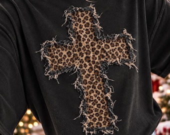 May include: A black short-sleeve t-shirt featuring a large cross applique on the chest. The cross is made of a brown and black leopard print fabric with frayed, distressed edges that create a textured, raw appearance against the solid black shirt.
