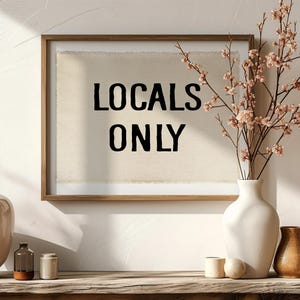 Pode incluir: Uma obra de arte emoldurada com o texto "LOCALS ONLY" em letras pretas. A obra de arte &eacute; exibida em uma superf&iacute;cie de madeira com vasos decorativos e um arranjo floral. A cena &eacute; banhada pela luz natural, criando um ambiente acolhedor e convidativo.