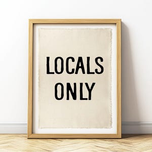 Pode incluir: Uma impress&atilde;o emoldurada com as palavras "LOCALS ONLY" em letras pretas. A impress&atilde;o tem um fundo bege e est&aacute; emoldurada em uma moldura de madeira marrom clara. A obra de arte &eacute; exibida em uma parede acima de um piso de madeira.