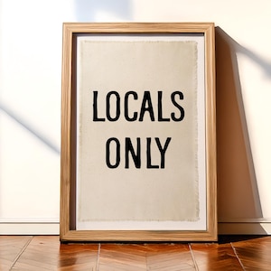 Pode incluir: Uma impress&atilde;o emoldurada com as palavras "LOCALS ONLY" em letras pretas em negrito. A impress&atilde;o tem um fundo texturizado esbranqui&ccedil;ado e est&aacute; emoldurada em uma moldura de madeira marrom clara. A obra de arte est&aacute; encostada em uma parede branca em um ch&atilde;o de madeira.
