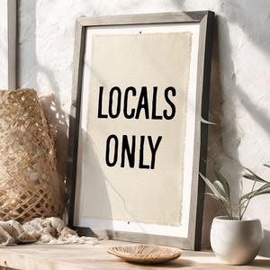 Pode incluir: Impress&atilde;o emoldurada com as palavras pretas "LOCALS ONLY" sobre um fundo bege. A moldura cinza est&aacute; sobre uma mesa de madeira, junto a uma cesta tecida, uma tigela pequena e uma planta em vaso, criando um ambiente r&uacute;stico.