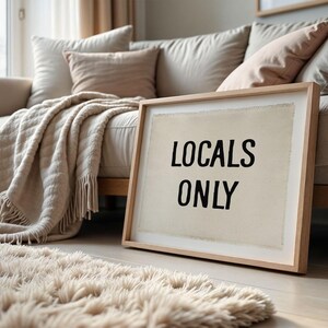 Pode incluir: Uma obra de arte emoldurada com o texto "LOCALS ONLY" em preto sobre um fundo creme, encostada a um sof&aacute; bege. Um tapete fofo e uma manta com franjas adicionam textura ao interior acolhedor.