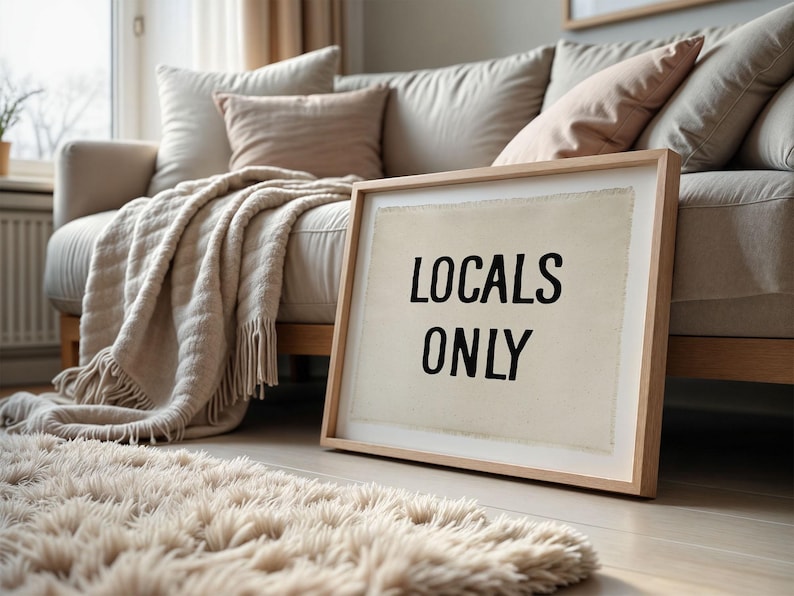 Pode incluir: Uma obra de arte emoldurada com o texto "LOCALS ONLY" em preto sobre um fundo creme, encostada a um sof&aacute; bege. Um tapete fofo e uma manta com franjas adicionam textura ao interior acolhedor.