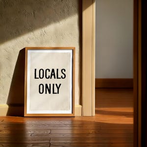 Pode incluir: Uma impress&atilde;o emoldurada com as palavras "LOCALS ONLY" em preto sobre um fundo creme. A moldura &eacute; de madeira castanha clara. A impress&atilde;o est&aacute; encostada a uma parede bege texturizada, com um ch&atilde;o de madeira em primeiro plano.