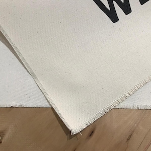 Pode incluir: Close-up de tecido de lona branco sujo com uma borda crua. O tecido tem uma trama vis&iacute;vel. A letra "W" &eacute; parcialmente vis&iacute;vel em preto na tela. O tecido &eacute; colocado sobre uma superf&iacute;cie de madeira.