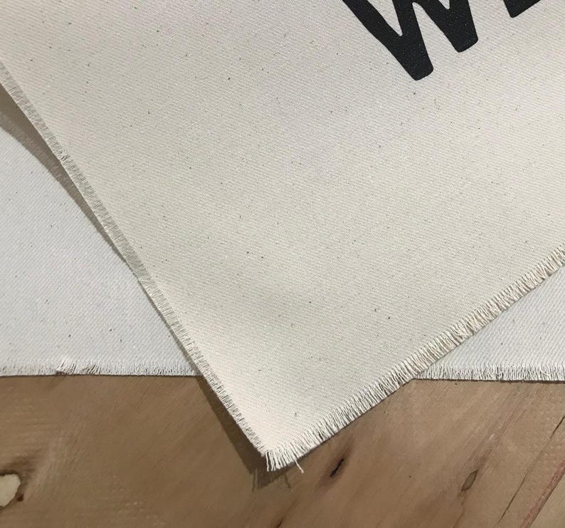 Pode incluir: Close-up de tecido de lona branco sujo com uma borda crua. O tecido tem uma trama vis&iacute;vel. A letra "W" &eacute; parcialmente vis&iacute;vel em preto na tela. O tecido &eacute; colocado sobre uma superf&iacute;cie de madeira.