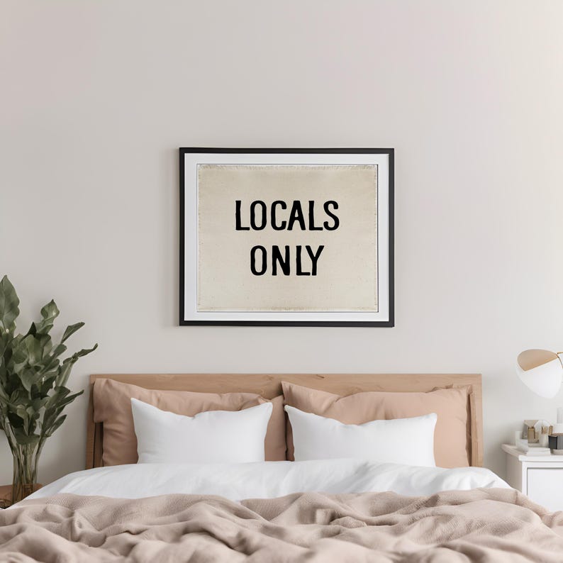 Pode incluir: Uma impress&atilde;o emoldurada com o texto "LOCALS ONLY" em preto, pendurada acima de uma cama. A cama tem travesseiros brancos, fronhas cor de p&ecirc;ssego e um cobertor bege. Um vaso de plantas est&aacute; &agrave; esquerda e uma mesa de cabeceira com uma l&acirc;mpada &agrave; direita.