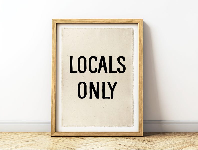 Pode incluir: Uma impress&atilde;o emoldurada com as palavras "LOCALS ONLY" em letras pretas. A impress&atilde;o tem um fundo bege e est&aacute; emoldurada em uma moldura de madeira marrom clara. A obra de arte &eacute; exibida em uma parede acima de um piso de madeira.