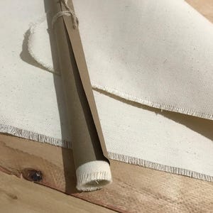 Pode incluir: Uma tela enrolada, embrulhada em papel castanho e amarrada com barbante, repousa sobre uma superf&iacute;cie de madeira ao lado de um grande peda&ccedil;o de tela. A tela tem uma borda crua e desfiada. O rolo de papel est&aacute; parcialmente desenrolado.