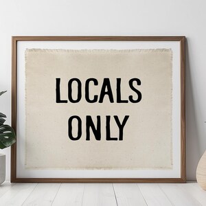 Pode incluir: Uma obra de arte emoldurada com o texto "LOCALS ONLY" em letras pretas sobre um fundo bege. A moldura &eacute; marrom. Duas plantas em vasos, uma com folhas verdes grandes e a outra com folhas verdes pontiagudas, ladeiam a obra de arte.