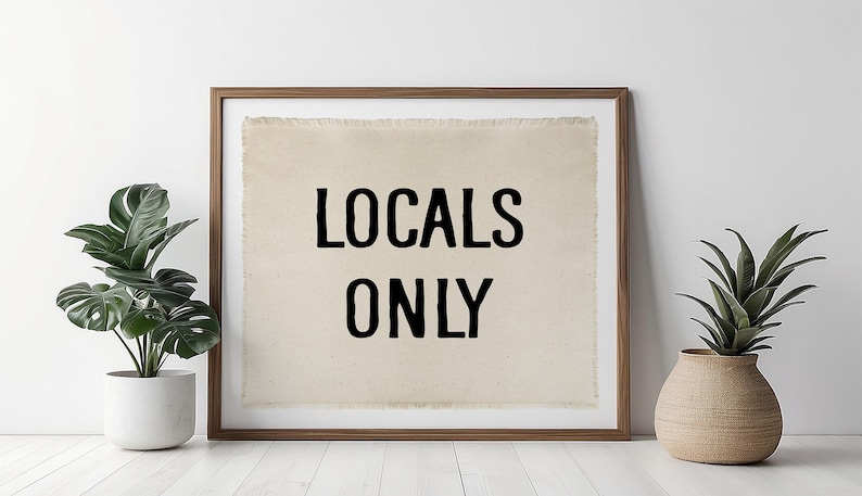 Pode incluir: Uma obra de arte emoldurada com o texto "LOCALS ONLY" em letras pretas sobre um fundo bege. A moldura &eacute; marrom. Duas plantas em vasos, uma com folhas verdes grandes e a outra com folhas verdes pontiagudas, ladeiam a obra de arte.