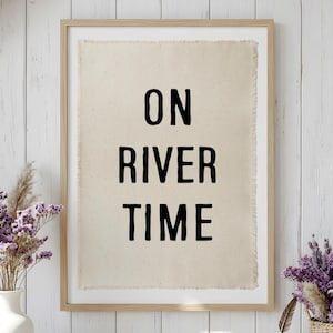 May include: A framed wall art piece featuring a rectangular piece of light beige fabric with frayed edges. The text ON RIVER TIME is printed in bold, black, sans-serif capital letters centred on the fabric within a light wood frame.