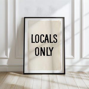 Pode incluir: Uma impress&atilde;o emoldurada com as palavras "LOCALS ONLY" em letras pretas em negrito. A impress&atilde;o tem um fundo texturizado esbranqui&ccedil;ado e est&aacute; emoldurada em uma moldura preta. A obra de arte est&aacute; encostada em uma parede branca em um ch&atilde;o de madeira.